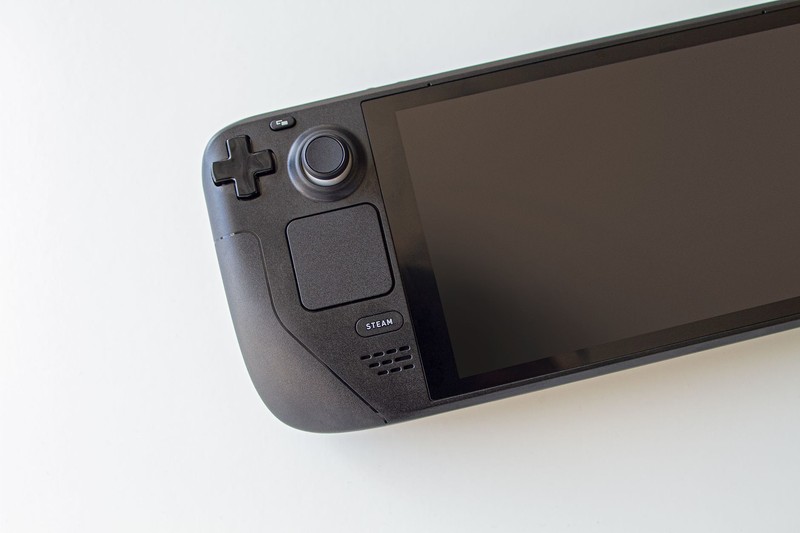 A Steam Deck handheld gaming console resting on a white surface, showing the Steam logo and control layout