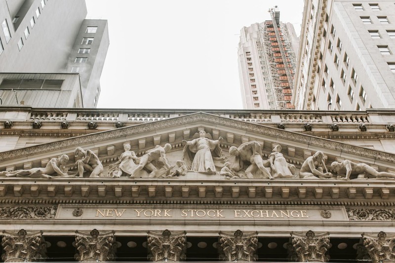 The neoclassical facade of the New York Stock Exchange building with columns, sculptural frieze, and the Manhattan financial district skyline behind it