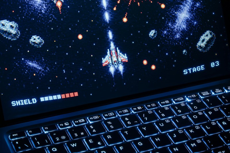 A space shooter video game displayed on a laptop screen with an illuminated keyboard below