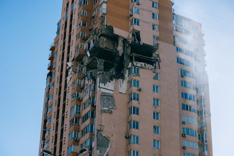 A multi-story residential apartment building with a large section of its facade destroyed, exposing interior structure and debris against a clear blue sky.