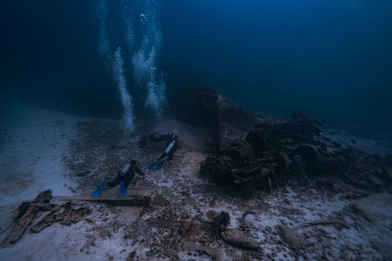 Two scuba divers explore the remains of a sunken shipwreck on the ocean floor, with bubbles rising toward the surface