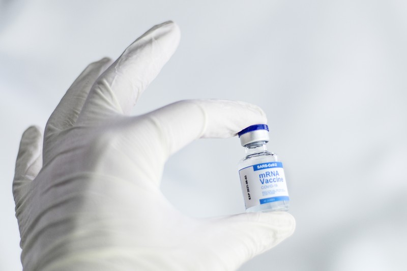 A gloved hand holds a small mRNA vaccine vial against a blurred light background
