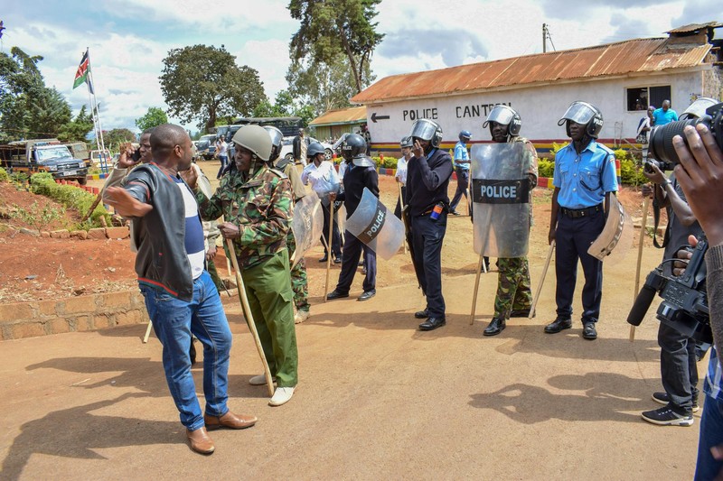 Police officers in riot gear face a civilian protester near a building flying a Kenyan flag